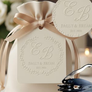 May include: A cream-colored gift bag with a satin ribbon and tag. The tag features embossed initials "EB", the names "Emily & Bryan", and the date "EST. 2026" within a floral wreath. A round tag with the same design is also visible.