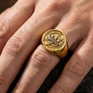 Könnte beinhalten: Ein goldfarbener Siegelring mit einem detaillierten Emblem mit Krone, Schwertern und Text. Der Ring ist rund mit flacher Oberseite und wird am Finger getragen. Der Ring besteht aus glänzendem, metallischem Material.
