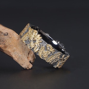 Puede incluir: Un anillo negro y dorado con un diseño floral detallado. El anillo está hecho de metal y tiene un acabado texturizado.