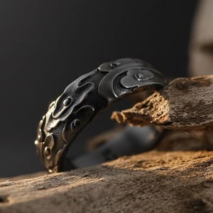 Può includere: Un anello grigio scuro con un design a nuvola ornato. L'anello è in metallo e ha una superficie dettagliata e strutturata. L'anello è esposto su un pezzo di legno, con uno sfondo scuro.