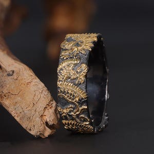 May include: A black and gold ring with an intricate design. The ring is made of metal and has a textured surface.