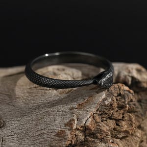 Puede incluir: Un anillo negro con un diseño de serpiente texturizado. El anillo está hecho de un metal oscuro y se encuentra sobre una superficie de madera rústica. La cabeza de la serpiente forma el extremo del anillo, creando un accesorio único y elegante.