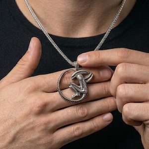 Op de afbeelding: Zilverkleurige hangerketting met een Alien-ontwerp. De hanger toont het wezen in een cirkelvormig frame, hangend aan een ketting. De ketting wordt gedragen over een zwart T-shirt.