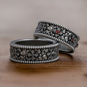 Può includere: Due anelli d'argento con disegni intricati e pietre preziose rosse. Gli anelli sono impilati uno sopra l'altro su una superficie di legno.