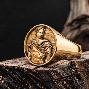 May include: A gold signet ring featuring a circular face with a raised relief portrait of a figure in classical attire. The ring has a polished, smooth band and a detailed, textured depiction of a person wearing a draped garment on the face.