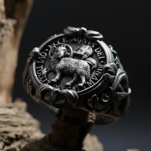 May include: A detailed silver ring featuring a central relief of a lamb within a circular frame. The ring has intricate detailing and text around the lamb, with a dark, antique finish. The ring is set against a textured wooden background.