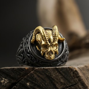 May include: A detailed, gothic-style ring featuring a gold-colored ram skull design set against a dark, engraved band. The skull has prominent horns and is the focal point of the ring. The ring is displayed on a piece of wood.
