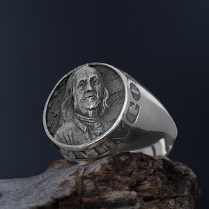 Könnte beinhalten: Ein silberner Ring mit einem detaillierten Porträt eines Mannes auf der Vorderseite des Rings. Der Ring liegt auf einem Stück verwittertem Holz.