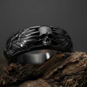 May include: A silver ring with a skull motif at the centre, surrounded by detailed wing designs. The ring's intricate features are set against a dark backdrop, emphasizing the skull and feather-like wings.