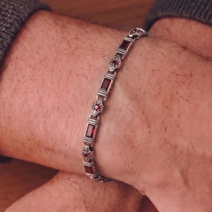 May include: A silver-toned metal bracelet featuring alternating rectangular and circular links. The rectangular links are set with deep red, baguette-cut stones, while the circular links contain small, round, faceted red stones.