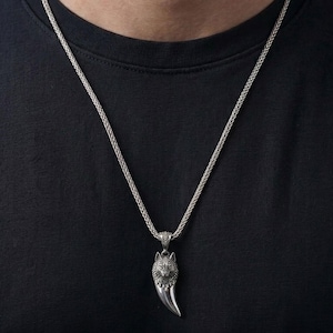 Op de afbeelding: Zilverkleurige ketting met een wolfskop-hanger en een klauwachtig ontwerp. De hanger hangt aan een gevlochten ketting. De ketting wordt gedragen op een zwart T-shirt.