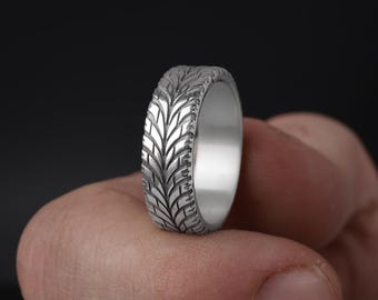 May include: A silver ring featuring a tyre tread design. The ring is held between fingers. The ring's surface has a detailed pattern, mimicking the tread of a tyre.