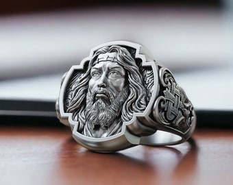 May include: A silver ring with a detailed design featuring a man's face in the center. The ring is set on a wooden surface.