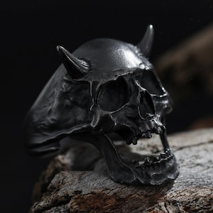 May include: A dark gray skull ring with curved horns. The ring has detailed facial features, including eye sockets, a nose cavity, and an open mouth. The ring is made of metal and sits on a piece of wood.