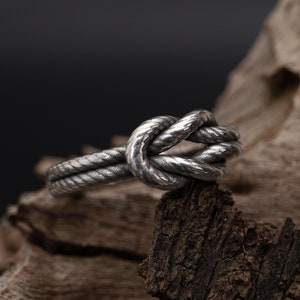 May include: A silver ring featuring a rope knot design. The ring is resting on a piece of weathered wood.
