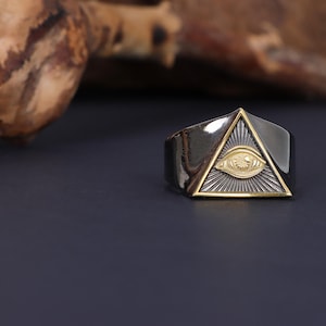 Può includere: Un anello nero e dorato con un design triangolare che presenta un simbolo di occhio onniveggente.