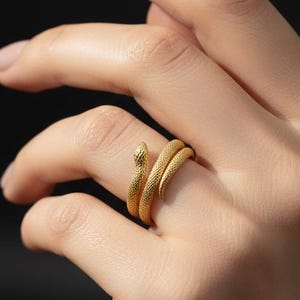 May include: A gold-coloured snake ring with textured scales wraps around a finger. The snake's head is raised, creating a unique design. The ring is set against a dark background.
