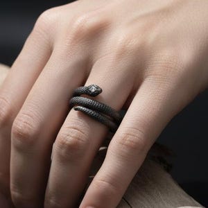May include: A dark grey snake ring with a coiled design. The ring has a small, clear stone at the snake's head. The ring is worn on a finger, with a wooden branch in the background.