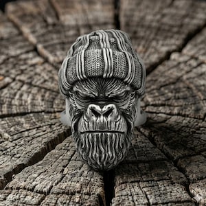 Könnte beinhalten: Ein silberner Ring mit einem detaillierten Gorillakopf, der eine Strickmütze trägt. Das Gesicht des Gorillas weist aufwendige Texturen auf, mit einer gerunzelten Stirn und einem Bart. Der Ring ist vor einem rustikalen Holzgrund.