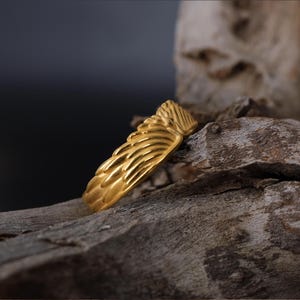 May include: A gold ring with a textured design resembling feathers. The ring is on a piece of weathered wood.