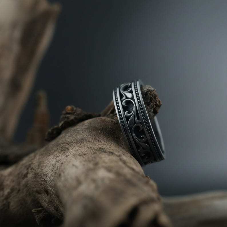 Puede incluir: Un anillo plateado con un intrincado dise&ntilde;o floral en espiral. El anillo se muestra sobre un fondo de madera natural, resaltando su dise&ntilde;o detallado. La superficie del anillo tiene una apariencia texturizada.