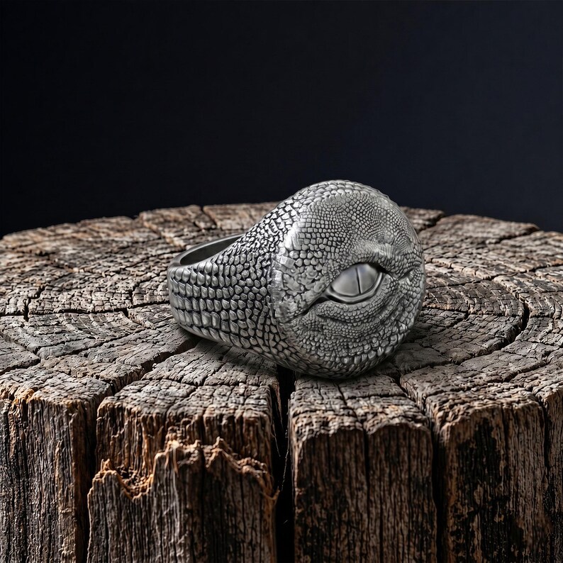 Puede incluir: Un anillo de tono plateado con un patr&oacute;n texturizado similar a escamas de reptil en toda la banda y un motivo de ojo central prominente con una pupila lisa en forma de almendra incrustada en la superficie met&aacute;lica detallada.