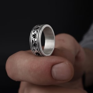 Puede incluir: Un anillo de plata con un dise&ntilde;o de volutas ornamentado y repetitivo. El anillo est&aacute; sostenido por el dedo de una persona. El dise&ntilde;o del anillo es negro y el anillo en s&iacute; es plateado. El anillo es de estilo banda.