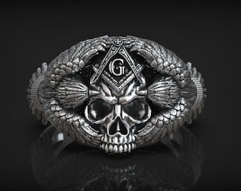 Puede incluir: Un anillo de plata con un diseño de calavera, rodeado de alas y un símbolo masónico con la letra 'G' en el centro.