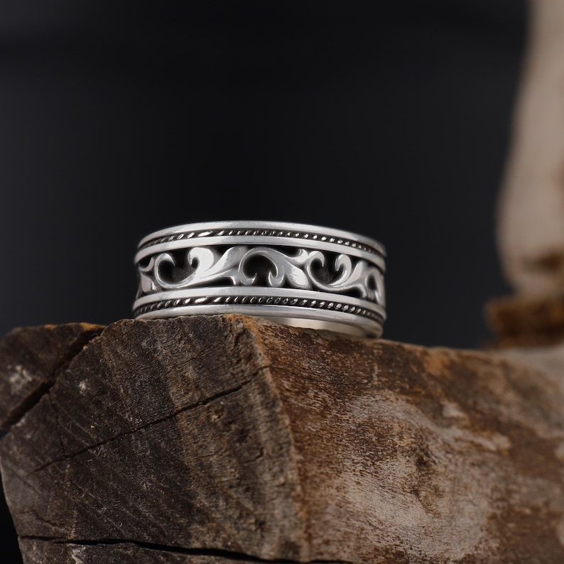 Puede incluir: Un anillo de plata con un dise&ntilde;o ornamental y ondulado. El anillo presenta un patr&oacute;n detallado de l&iacute;neas y curvas arremolinadas, con un borde de cuentas. El anillo se muestra sobre un trozo de madera sobre un fondo oscuro, resaltando el intrincado trabajo en metal.