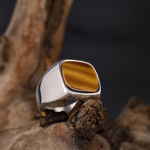 May include: A silver ring featuring a square tiger's eye gemstone in shades of brown and gold. The ring is displayed on a piece of wood, highlighting the contrast between the polished metal and the natural wood grain.