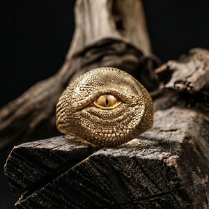Puede incluir: Un anillo de tono dorado con un patr&oacute;n texturizado de escamas de reptil en toda su superficie. El centro del anillo muestra un ojo realista prominente con una pupila vertical y un iris amarillo brillante, incrustado en las escamas.