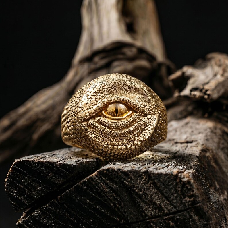 Puede incluir: Un anillo de tono dorado con un patr&oacute;n texturizado de escamas de reptil en toda su superficie. El centro del anillo muestra un ojo realista prominente con una pupila vertical y un iris amarillo brillante, incrustado en las escamas.
