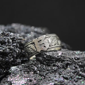 Pu&ograve; includere: Un anello d'argento con un ornato motivo floreale e a lettere. L'anello &egrave; appoggiato su una superficie scura e strutturata. La lettera "B" &egrave; ben visibile al centro dell'anello.