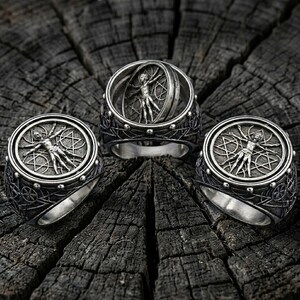May include: Three silver rings with intricate designs. Each ring features a detailed depiction of the Vitruvian Man within a circular frame. The rings are set against a dark, textured wooden surface, highlighting the metallic sheen and detailed craftsmanship.