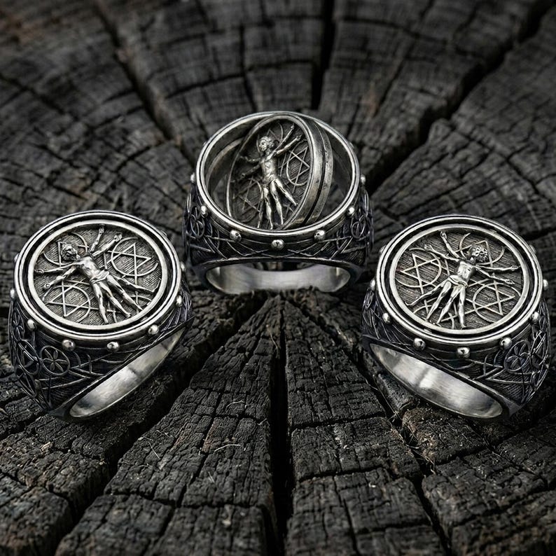 May include: Three silver rings with intricate designs. Each ring features a detailed depiction of the Vitruvian Man within a circular frame. The rings are set against a dark, textured wooden surface, highlighting the metallic sheen and detailed craftsmanship.