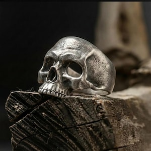 May include: A silver-colored skull ring with detailed facial features, including eye sockets, a nose cavity, and teeth. The ring is resting on a piece of weathered wood, creating a contrast between the metallic ring and the natural texture of the wood.