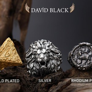 May include: Three detailed rings: a gold-plated pyramid, a silver lion head, and a rhodium-plated ring with a lamb emblem. The rings are displayed on a textured wooden surface, with the brand name "DAVID BLACK" at the top.