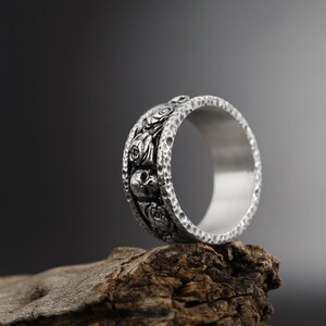 K&ouml;nnte beinhalten: Ein silberner Ring mit einem detaillierten Design aus Totenk&ouml;pfen und Rosen. Der Ring liegt auf einem St&uuml;ck verwittertem Holz.