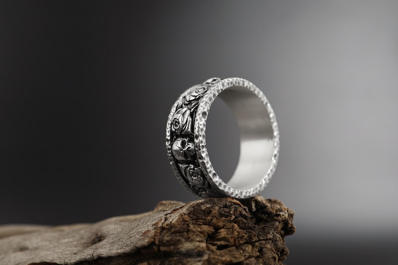 K&ouml;nnte beinhalten: Ein silberner Ring mit einem detaillierten Design aus Totenk&ouml;pfen und Rosen. Der Ring liegt auf einem St&uuml;ck verwittertem Holz.