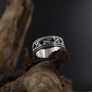 Puede incluir: Un anillo de plata con un dise&ntilde;o ornamental y ondulado. El anillo presenta un patr&oacute;n detallado de l&iacute;neas y curvas fluidas, con un fondo oscuro. El anillo se exhibe sobre un trozo de madera.
