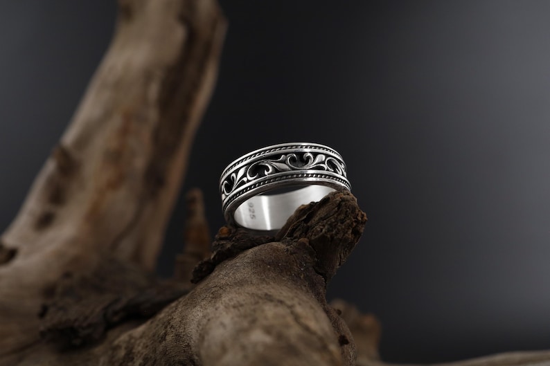 Puede incluir: Un anillo de plata con un dise&ntilde;o ornamental y ondulado. El anillo presenta un patr&oacute;n detallado de l&iacute;neas y curvas fluidas, con un fondo oscuro. El anillo se exhibe sobre un trozo de madera.