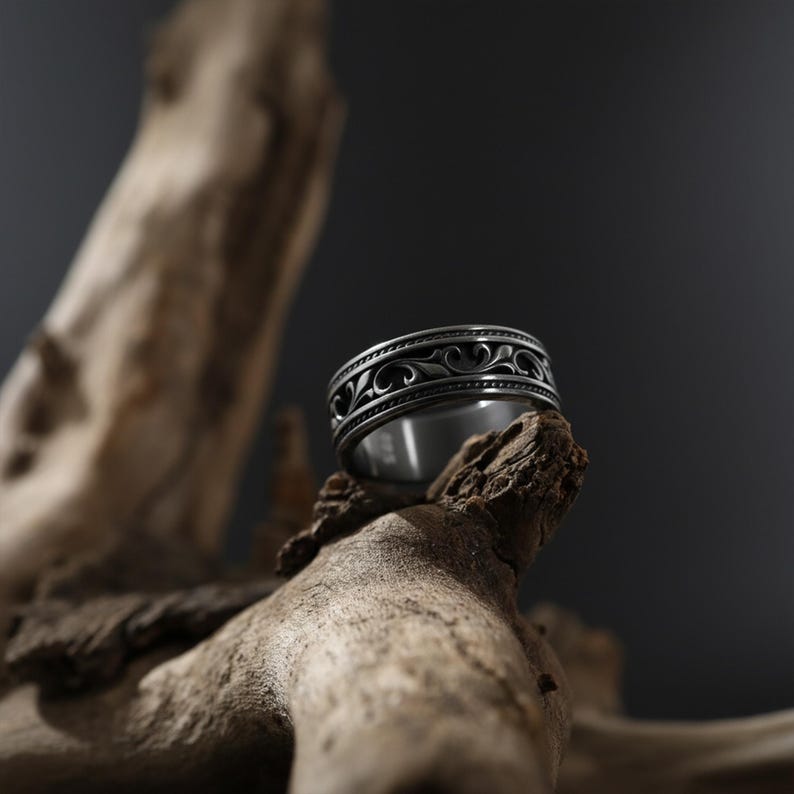 Puede incluir: Un anillo plateado con un intrincado dise&ntilde;o de remolinos alrededor de la banda. El anillo se exhibe sobre un trozo de madera envejecida contra un fondo oscuro, resaltando su dise&ntilde;o detallado y brillo met&aacute;lico.