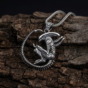 May include: A silver-colored pendant necklace featuring a detailed depiction of an alien creature. The alien is seated within a circular frame, with a textured chain. The pendant is set against a dark, textured wooden surface.