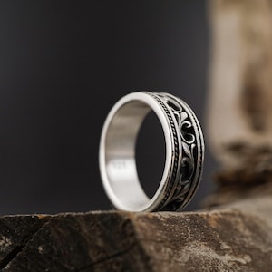 Puede incluir: Un anillo de plata con un dise&ntilde;o floral ornamentado y de color oscuro. El anillo se encuentra sobre un fondo oscuro y descansa sobre un trozo de madera. La banda del anillo presenta un patr&oacute;n detallado y el metal tiene un acabado pulido.