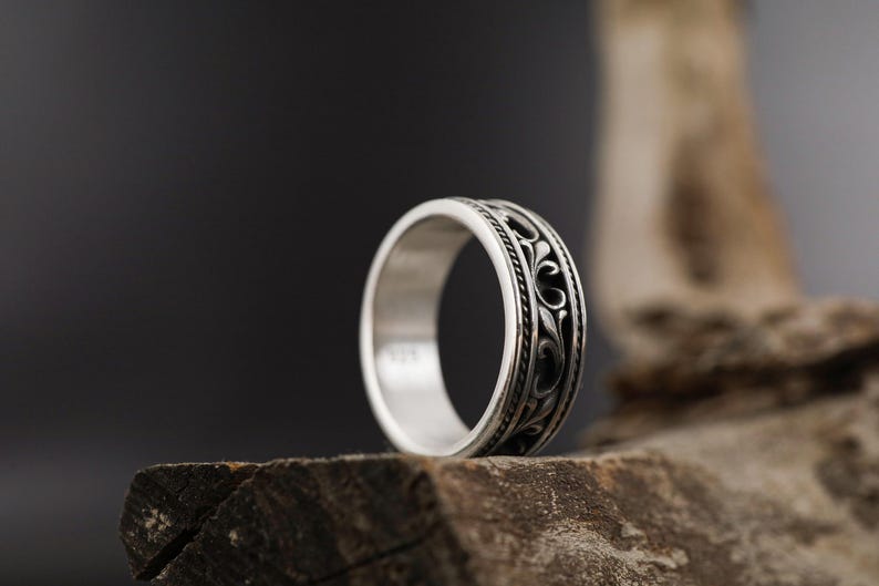 Puede incluir: Un anillo de plata con un dise&ntilde;o floral ornamentado y de color oscuro. El anillo se encuentra sobre un fondo oscuro y descansa sobre un trozo de madera. La banda del anillo presenta un patr&oacute;n detallado y el metal tiene un acabado pulido.