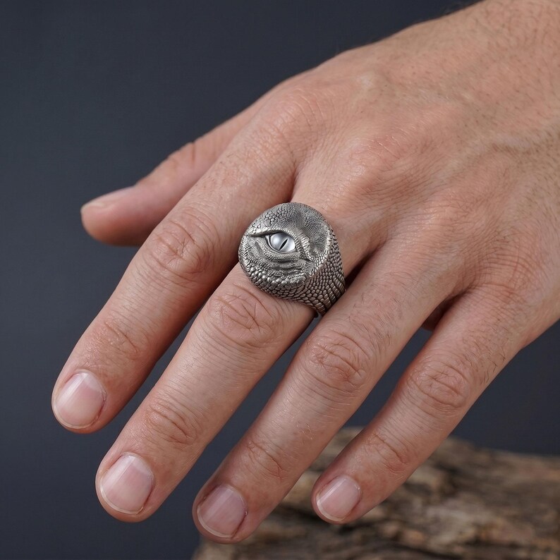 Puede incluir: Un anillo de tono plateado con un patr&oacute;n detallado de piel de reptil texturizada que rodea un ojo ovalado central, liso y de color claro. La superficie met&aacute;lica tiene un acabado oscuro y oxidado en los huecos para resaltar los detalles.