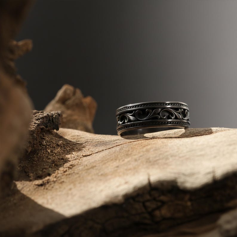 Puede incluir: Un anillo gris oscuro con un intrincado dise&ntilde;o floral. El anillo se encuentra sobre una superficie de madera natural, con un fondo borroso. El dise&ntilde;o del anillo presenta un motivo repetitivo en forma de voluta, creando un aspecto cl&aacute;sico y elegante.