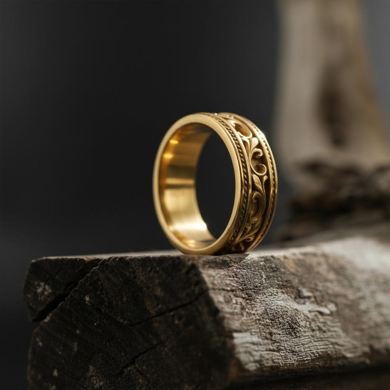 Puede incluir: Un anillo dorado con un dise&ntilde;o grabado y ornamentado. El anillo presenta un patr&oacute;n floral repetido alrededor de la banda. El anillo se exhibe sobre un trozo de madera envejecida, con un fondo oscuro.