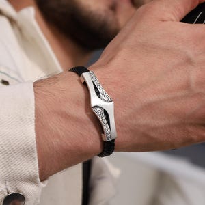 May include: A black leather bracelet with a silver clasp. The clasp has an intricate, etched design. The bracelet is worn on a wrist, and the person is wearing a white jacket.