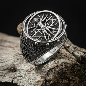 May include: A silver ring featuring a detailed depiction of the Vitruvian Man within a circular frame. The ring has intricate geometric patterns and the inscription ".925" on the band, indicating its silver composition. The ring is displayed on a piece of wood.